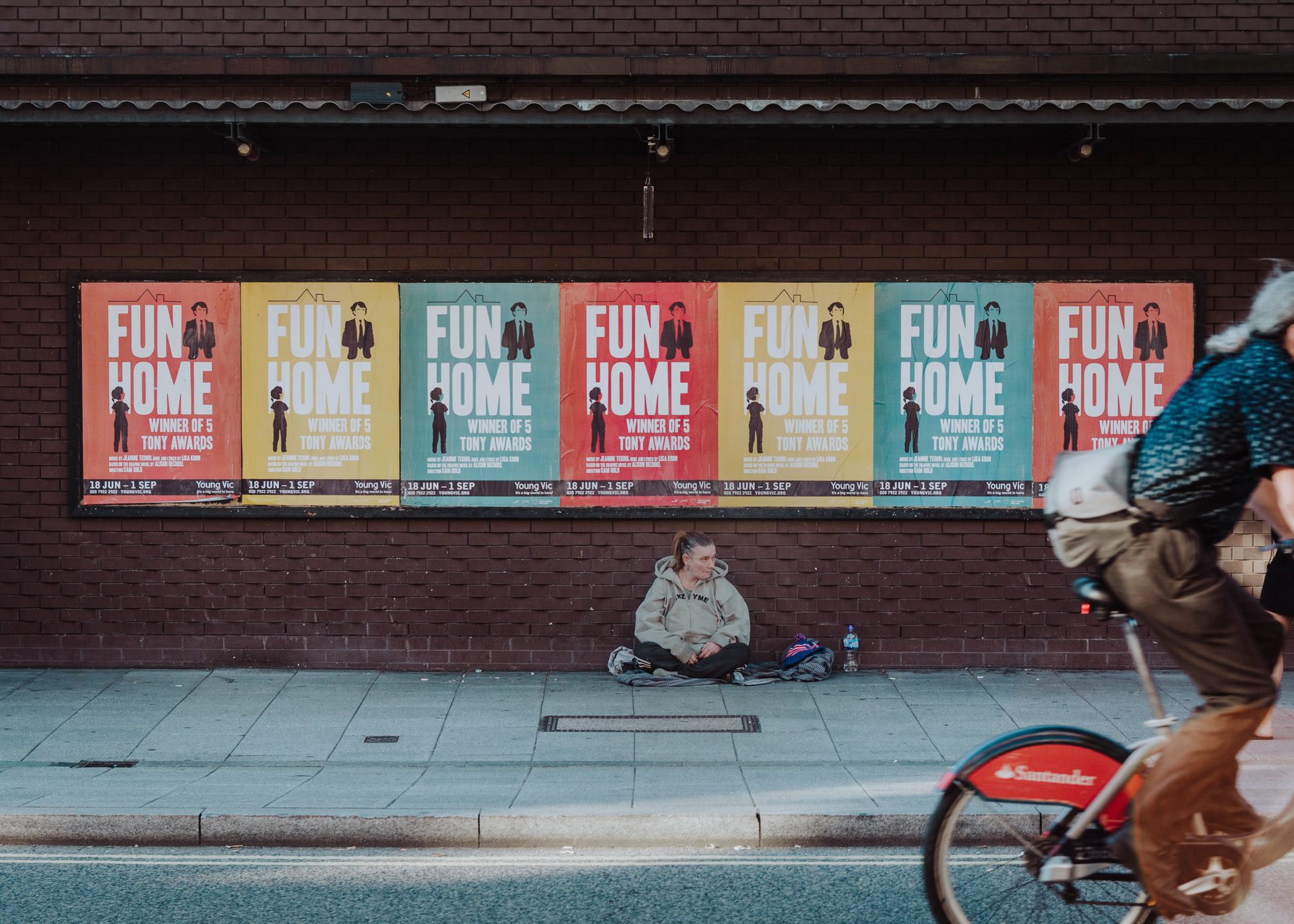 Homeless woman sitting in front of colorful advertisement for Fun Home